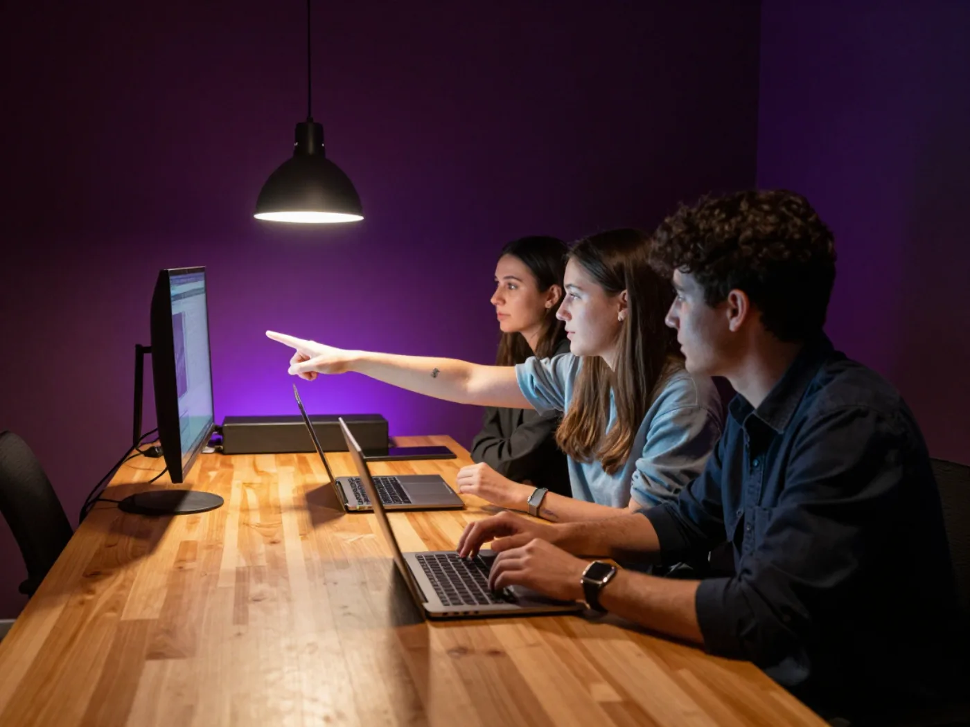 Trzy osoby pracujące przy laptopach w niewielkim studiu oświetlonym fioletowym światłem